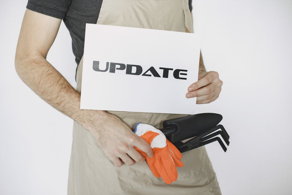 A young man with dark curly hair, wearing a navy blue t-shirt, beige shorts, white sneakers, and orange work gloves, is engaging in garden maintenance outdoors. He is using a long-handled shovel to di
